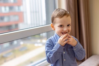 Pencerenin yanında mavi gömlekli neşeli küçük bir çocuk, şakacı bir şekilde dilini çıkarıp gülümsüyor. Metin için boşluk