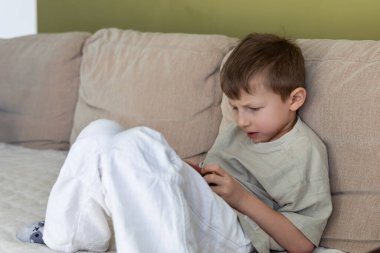 Genç çocuk akıllı telefon ekranına odaklanmış. Genç bir çocuk bej bir kanepede oturur, dikkatle akıllı telefonuna bakar, bir oyuna veya evdeki dijital içeriğe derinden bağlıdır.