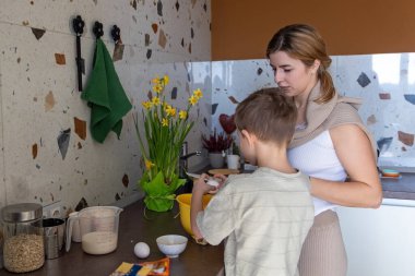 Anne, oğlunun mutfakta yumurta kırmasına yardım ediyor. Bir anne, genç oğlunun modern bir mutfakta birlikte yemek pişirirken yumurta kırmasını izler. Öğrenme ve bağlanmayı teşvik eder.