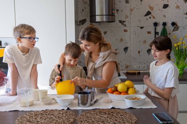 Anne ve çocuklar mutfakta birlikte yemek pişiriyorlar. Bir anne ve üç çocuğu modern bir mutfakta birlikte yemek pişirmeyi, bir kasede malzemeleri karıştırmayı ve aile olarak kaynaşmayı seviyorlar.