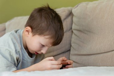 Genç çocuk akıllı telefon ekranına odaklanmış. Kanepede otururken akıllı telefona yoğun bir şekilde odaklanan genç bir çocuk ekran bağımlılığını, konsantrasyonunu ve olası miyop riskini vurguluyor.