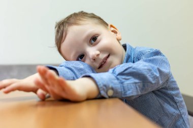 Masaya yaslanan mutlu genç çocuk. Mavi kareli gömlekli neşeli genç bir çocuk ahşap bir masaya yaslanıyor, gülümsüyor ve elleriyle elini uzatıyor.