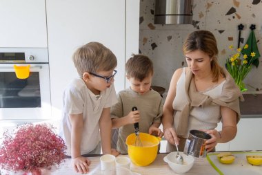 Anne ve Oğulları Mutfakta Birlikte Yemek Yapıyor. Bir anne ve iki küçük oğlu, parlak ve modern bir mutfakta birlikte yemek pişirirken sarı bir kâsede malzemeleri karıştırıyorlar.