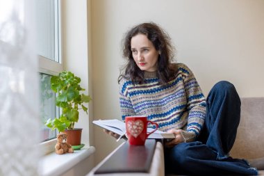 Pencerenin yanında kahve okuyan bir kadın. Rahat bir kazak giymiş bir kadın pencerenin kenarında oturur, yanında kırmızı kahve fincanı olan bir kitap okur, huzurlu bir anın tadını çıkarır.