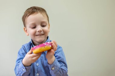 Mutlu çocuk pembe bir donuta hayran. Mavi tişörtlü neşeli genç bir çocuk heyecanlı bir şekilde iki eliyle tutarak serpiştirilmiş pembe bir donuta bakar. Metin için boşluk