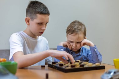 Evde dama oynayan iki kardeş. Beyaz gömlekli büyük bir çocuk ve mavi gömlekli küçük bir çocuk masada dama oynuyorlar, genç görünümlü hayal kırıklığına uğramış.