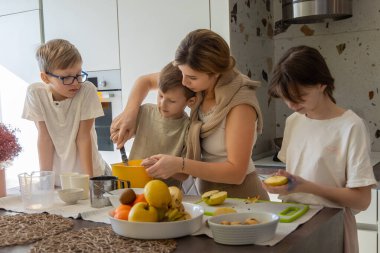 Anne ve çocuklar mutfakta birlikte yemek pişiriyorlar. Bir anne ve üç çocuğu parlak bir mutfakta birlikte yemek hazırlıyor, malzemeleri karıştırıyor ve aile zamanlarının tadını çıkarırken meyveleri kesiyorlar.
