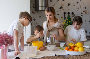 Anne ve çocuklar mutfakta birlikte yemek pişiriyorlar. Bir anne ve üç çocuğu sıcak bir mutfakta birlikte yemek hazırlıyor, malzemeleri karıştırıyor ve aile zamanının tadını çıkarırken meyve kesiyorlar..