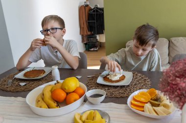 Kahvaltıda meyveli krep yiyen iki çocuk. İki genç çocuk, ev ortamında sıcak bir yemek masasında krep, krema ve taze meyve ile ev yapımı kahvaltıdan zevk alıyor.