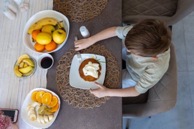 Çocuk kahvaltıda krep ve meyveyle eğleniyor. Genç bir çocuk yemek masasında kremayla kaplı bir tabak kremayla oturuyor. Taze meyvelerle çevrili.