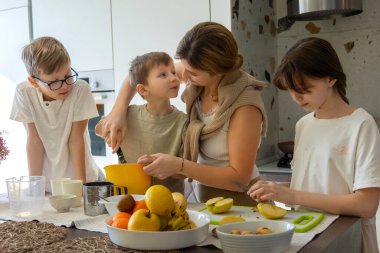Anne evde çocuklarla yemek pişiriyor. Bir anne ve üç çocuğu sıcak bir mutfakta birlikte yemek hazırlıyorlar. Meyve kesiyorlar ve yemek için malzemeleri karıştırıyorlar.