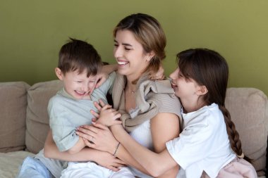 Mutlu anne kucaklaması ve çocuklarla oynaması. Neşeli anne oğlunu ve kızını rahat bir kanepede kucaklarken gülüyor. Mutluluk ve sıcaklıkla dolu sevgi dolu bir aile anı.