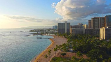 Tropical Beach ve City Skyline 'ın havadan görüntüsü. Güneş batarken sakin okyanus sularına bakan altın kumu, palmiyeleri ve yüksek binaları olan çarpıcı bir tropikal sahil manzarası.