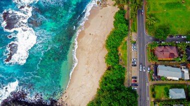 Tropik Plaj ve Sahil Yolu 'nun havadan görünüşü. Turkuaz okyanus dalgaları, yemyeşil yemyeşil ve park halindeki arabalarla dolu sahil yolunun çarpıcı bir görüntüsü.