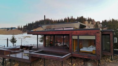 Karlı dağ manzarasında modern ahşap kulübe. Çağdaş ahşap kulübe, karlı bir dağlık alana yerleştirilmiş büyük camdan pencereler, rahat bir geri çekilme ve manzaralı orman manzarası sunuyor..