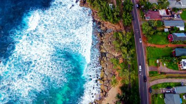 Kıyı yolu ve okyanus dalgalarının havadan görünüşü. Yukarıdan aşağıya insansız hava aracı görüntüleri okyanus dalgaları, kayalık kıyılar ve evlerle ve yeşilliklerle kaplı bir yol ile dolu canlı bir kıyı şeridi gösteriyor.