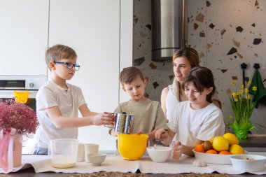 Modern mutfakta birlikte yemek pişiren mutlu bir aile. Üç çocuk ve bir kadın, neşeli bir atmosferde kaseler, meyveler ve çiçeklerle çevrili, aydınlık ve modern bir mutfakta yemek pişirmekten hoşlanırlar..
