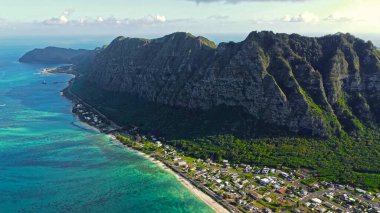 Engebeli Hawaii Kayalıklarının Altındaki Sahil Kasabası. Berrak turkuaz Hawaii kıyı şeridi boyunca sarp yeşil uçurumların dibinde yuva yapmış tropikal bir sahil kasabasının hava manzarası.