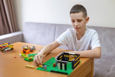 Çocuk renkli plastik yapı tuğlalarıyla oynuyor. Beyaz gömlekli genç bir masada oturuyor, renkli plastik tuğlalarla yapıları birleştiriyor, yaratıcı yapı oyunlarına odaklanıyor.