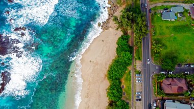 Turkuaz okyanus dalgalarının kumlu sahile düşüşünün havadan görüntüsü. Yeşil ağaçlarla kaplı kumlu bir sahile çarpan temiz turkuaz okyanus dalgalarının yukarıdan aşağı görüntüsü ve park edilmiş yakın bir yol.