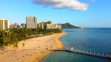 Waikiki Sahili ve Diamond Head 'in altın saatinde. Palmiye ağaçlarıyla Waikiki Sahili 'nin havadan görünüşü, kumlu sahildeki insanlar, mavi okyanus suları, şehir binaları, ve arka planda Diamond Head krateri.