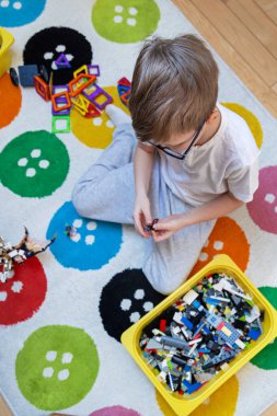 Çocuk renkli halıda yapı taşlarıyla oynuyor. Gözlük takan ve günlük kıyafetler giyen bir çocuk renkli bir halıya oturmuş düğmelerinde desenler, yapı taşları ve manyetik fayanslarla oynuyor..