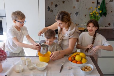 Anne çocuklarla aydınlık mutfakta yemek pişiriyor. Anne ve üç çocuk, meyve ve erzakla dolu sıcacık, aydınlık bir mutfakta malzemeleri hazırlayıp birlikte pişiriyorlar.
