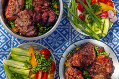 Piknik masasında ızgara et ve taze sebzelerin yakın çekimi. Güneşli bir açık hava pikniği sırasında mavi desenli masa örtüsünde servis edilen sulu ızgara et ve canlı taze sebzelerin ayrıntılı görüntüsü.