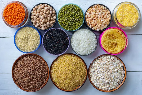 Beyaz Tahta Masa 'da Tahıl, Legumes ve Makarna. Beyaz ahşap bir yüzey üzerinde renkli kâselerde sergilenen çeşitli tahıllar, baklagiller ve makarna.