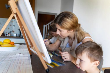 Anne, çocuklarla mutfak masasında resim yapıyor. Anne ve kızı masadaki resim projesine odaklanırken, genç bir çocuk da neşeli bir somurtkanlıkla bakıyor.