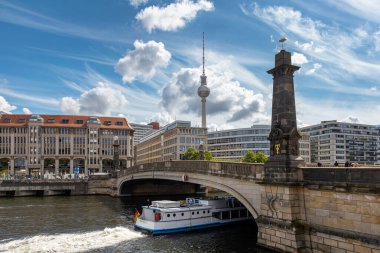 Berlin, 26.05.2025, Berlin Nehri Sahnesi TV Kulesi ve Köprüsü. Berlin 'deki River Spree' de taştan bir köprünün altından geçen tur teknesinin arkasında ikonik televizyon kulesi ve modern binalar var.