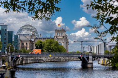 Almanya, Berlin, 26.05.2025, Berlin 'deki nehir manzaralı Reichstag binası. Berlins Reichstag 'ın cam kubbesi, ağaçlar ve modern binalar tarafından çerçevelenmiş, Spree Nehri boyunca görülüyor.