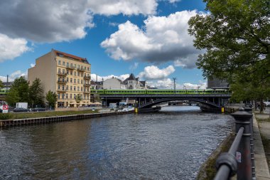 Almanya, Berlin, 26.05.2025, Berlin 'de Spree Nehri üzerindeki yeşil tren köprüsü. Berlin 'deki Spree Nehri üzerinde tarihi binalar ve dramatik bulutlarla çevrili metal bir köprüden geçen parlak yeşil bir tren görülüyor..