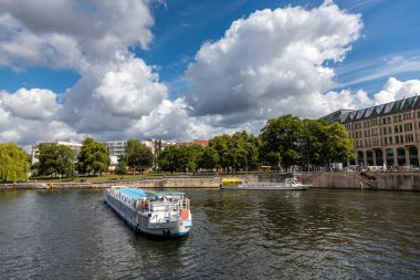 Berlin, 26.05.2025, Berlin River Spree 'deki Turist Gemisi. Berlin 'de Spree nehri boyunca ağaçlar ve tarihi binalarla dolu dramatik bir gökyüzü altında gezinen bir tekne.
