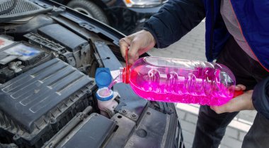 Arabanın ön camını dolduran adam yıkama sıvısı. Güneşli bir günde, parlak pembe bir ön cam temizleme sıvısını araba motorunun deposuna döken birinin yakın çekimi.