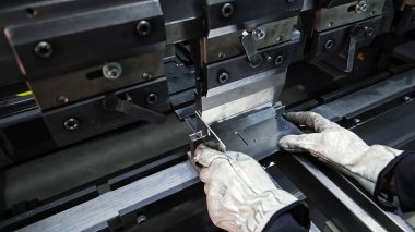 Metal levhayı bükmek için endüstriyel baskı freni kullanan bir işçi. Koruyucu eldiven giyen bir kişi düz bir metal levhayı doğru bir şekilde bükmek için fren makinası kullanıyor, ekipman mekanik bileşenleri açıkça görülüyor.
