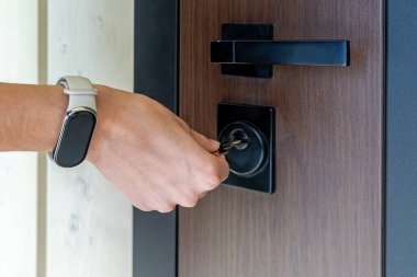 Anahtarı modern kapı kilidine yerleştiriyorum. Zindelik takip cihazı takan birinin yakın çekimi, siyah bir kilide anahtar takarak çağdaş ahşap bir kapının kilidini açmak.