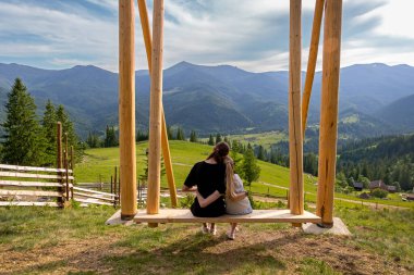 Kız kardeşler dağ manzaralı salıncakta oturuyorlar. İki kız kardeş, güneşli bir günde büyük bir ahşap salıncakta oturup huzurlu bir dağ manzarasının tadını çıkarırlar.