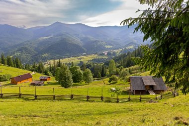 Kırsal ahşap evleri olan manzaralı dağ vadisi. Ahşap çiftlik binaları ve kırsal bir çit yemyeşil bir yamaçta, ağaçlarla kaplı, güneşli bir vadiye bakan ve parçalı bulutlu bir gökyüzünün altında orman dağları.