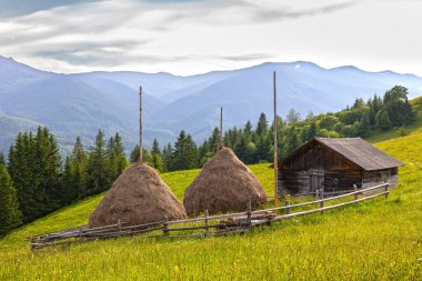 Mountain Meadow 'da saman yığınlı kırsal ahır. Geleneksel ahşap ahır ve arka planda sisli mavi dağ silsilesi ve orman yamacında iki saman yığını.