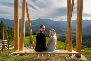 Sallanan İki Kız Dağlara Bakıyor. İki kız büyük bir salıncakta otururken gülümsüyor yeşil tepelerin, ormanın ve uzak dağ zirvelerinin manzarasına bakıyor.