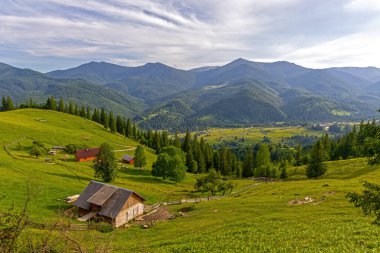 Ahşap ahırlı Dağ Köyü Manzarası. Yeşil tepelere dağılmış ahşap çiftlik binaları ve parçalı bulutlu bir gökyüzünün altında orman dağlarıyla huzurlu kırsal bir manzara.