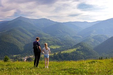 Baba ve kızı Dağ Vadisi Manzarası 'na hayran. Çimenli bir tepenin üzerinde duran erkek ve genç kız engin yeşil bir dağ manzarasına ve bulutlu mavi gökyüzünün altındaki orman vadisine bakıyor.