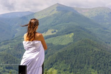 Dağlara bakan battaniyeye sarılı bir kadın. Rüzgarlı saçları olan bir kadın battaniyeye sarılmış, yeşil ormanlarla kaplı engin bir dağ manzarasına bakıyor.