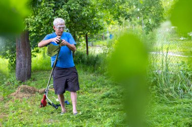 Kıdemli adam arka bahçede çim biçme makinesini koruyor. Koruyucu gözlük takan yaşlı bir adam yemyeşil bahçe düzeninde çalışırken çim biçme makinesini temizler ve inceler.