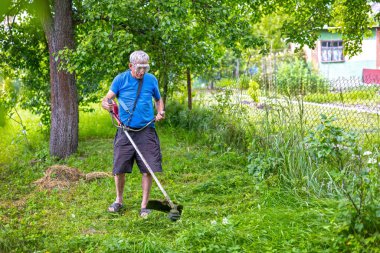 Yaşlı bir adam çim biçme makinesiyle çimleri buduyor. Gözlük takan son sınıf öğrencisi, bir ev ve çitin yanındaki yeşil bahçede otları biçmek için çim biçme makinesi kullanıyor.