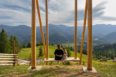 Dağ manzaralı iki kız kardeş salıncakta oturuyor. İki genç kız kardeş büyük bir salıncakta birlikte oturup, yeşil orman dağlarının güzel panoramik manzarasının tadını çıkarıyorlar.