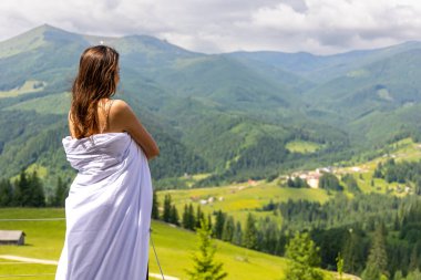 Beyaz çarşafa sarılı hamile bir kadın dağ vadisine bakıyor. Beyaz bir çarşafa sarılmış bir kadın, balkonda dikiliyor, engin yeşil bir dağ manzarasına ve aşağıdaki köye dikkatlice bakıyor..