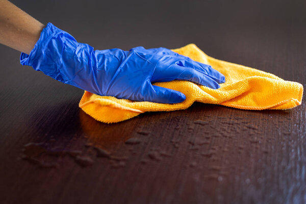 Рука в перчатках, вытирая влажную поверхность тканью. Крупный план человека в синей перчатке с использованием желтой ткани из микроволокна для очистки воды от темной деревянной поверхности
