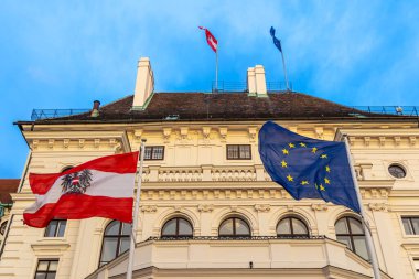 Avusturya ve AB Bayrakları Tarihi Binada Dalgalanıyor. Avrupa Birliği ve Avusturya bayrakları, ulusal birliği simgeleyen parlak mavi gökyüzünün altında büyük tarihi bir binanın önünde dalgalanıyor.